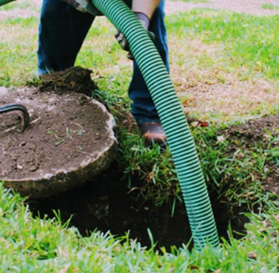 A green suction hose running down into an opened septic tank, the concrete lid levered aside on the grass